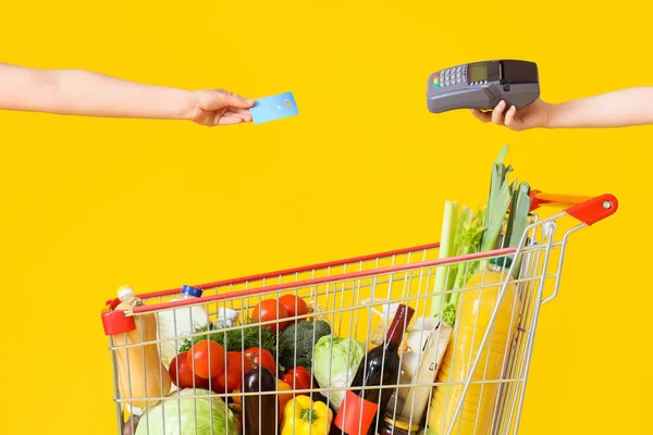 Female hands with credit card, payment terminal and shopping cart full of different fresh products on yellow background