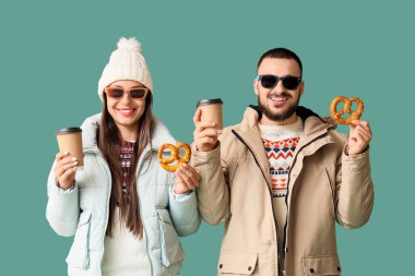 Young happy couple in winter clothes with tasty pretzels and cups of coffee on green background