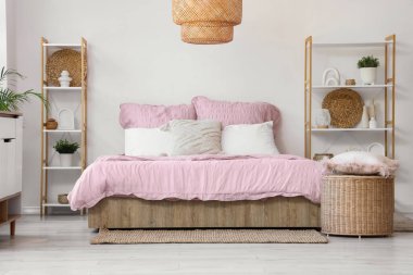 Bed with stylish pink bedding, shelving units, commode, wicker table and houseplants in bedroom