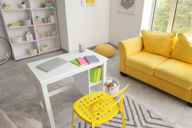Interior of room with shelving units, yellow sofa, laptop and notebooks on children's workplace, top view