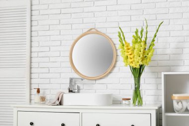 Glass vase with beautiful yellow gladiolus flowers on chest of drawers near white brick wall in bathroom