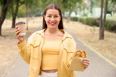 Young happy woman with tasty pretzel and cup of coffee in park
