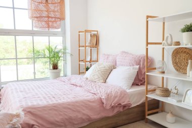 Bed with stylish pink bedding, shelving units and houseplants near window in bedroom