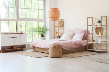 Bed with stylish pink bedding, shelving units, commode, wicker table and houseplants in bedroom