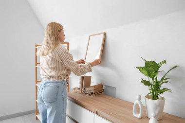 Beautiful young female interior designer hanging frame on white wall at home