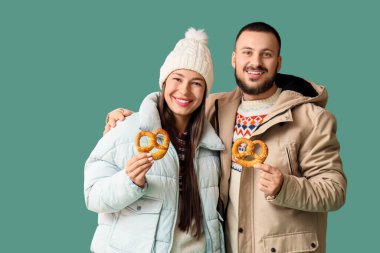 Young happy couple in winter clothes with tasty pretzels on green background