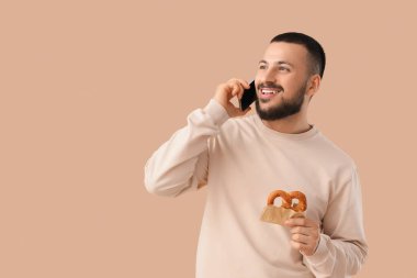 Young happy man with tasty pretzel talking by mobile phone on beige background