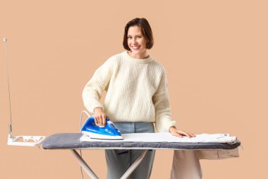 Bej arka planda giysileri ütüleyen genç bir kadın.