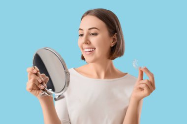 Young woman with occlusal splint and mirror on blue background, closeup
