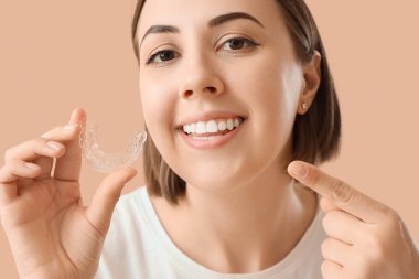 Young woman with occlusal splint pointing at her teeth on beige background