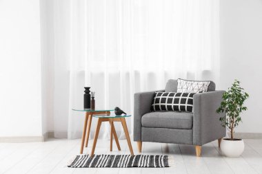 Interior of light living room with grey armchair, tables and window