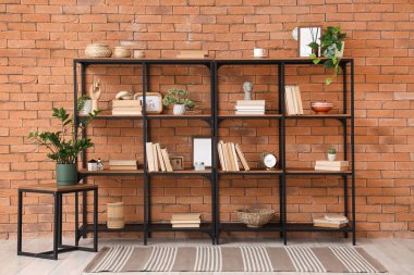 Shelf unit with books, decor and plants near brick wall in room