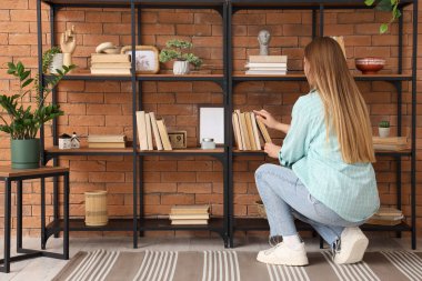 Young woman taking book from shelf in room, back view