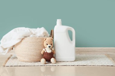 Yeşil duvarın yanındaki halının üzerinde oyuncak ayı ve deterjan olan çamaşır sepeti.