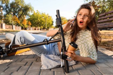 Young shocked woman with cup of coffee fallen off electric kick scooter in park