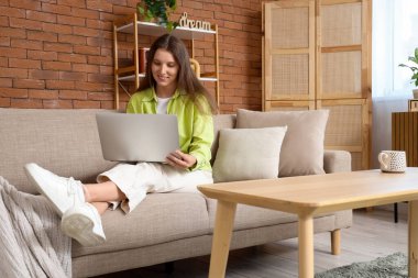 Evdeki koltukta modern laptop kullanan genç bir kadın.