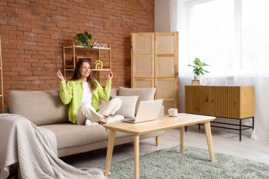 Dizüstü bilgisayarı olan genç bir kadın evdeki kanepede meditasyon yapıyor.