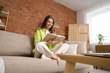 Evdeki kanepede kitap okuyan genç bir kadın.