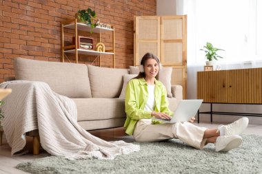 Kulaklıklı genç bir kadın evde dizüstü bilgisayar kullanıyor.