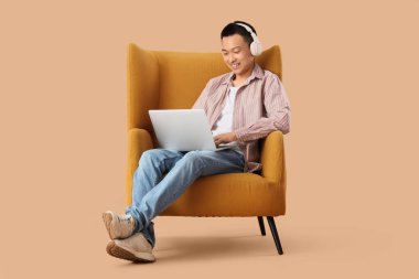 Young Asian man with headphones using laptop in armchair on beige background