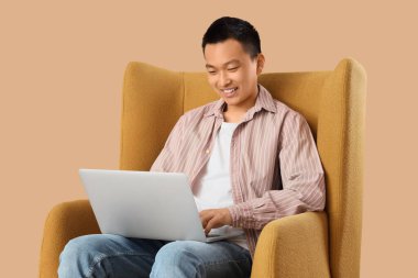 Young Asian man using laptop in armchair on beige background