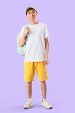 Male Asian student in cap with backpack on lilac background