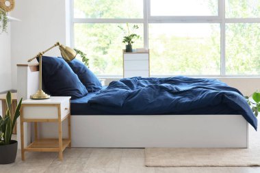 Bed with stylish blue bedding, commode, bedside table and houseplants near window in bedroom