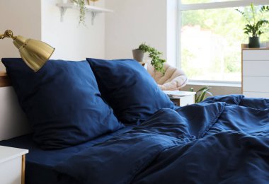 Bed with stylish blue bedding, commode and houseplants near window in bedroom