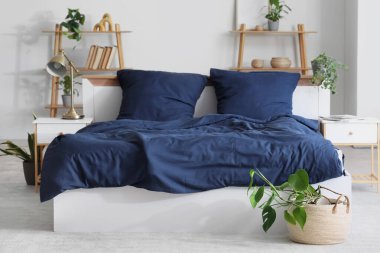 Bed with stylish blue bedding, lamp, shelving units, bedside tables and houseplants in bedroom