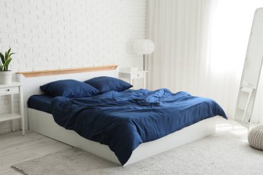 Bed with stylish blue bedding, bedside tables, mirror and lamp near white brick wall in bedroom