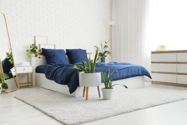 Bed with stylish blue bedding, commode, mirror, bedside table and houseplants near white brick wall in bedroom