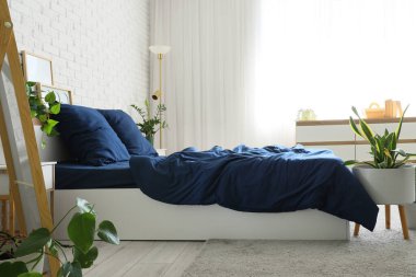Bed with stylish blue bedding, commode, lamp and houseplants near white brick wall in bedroom