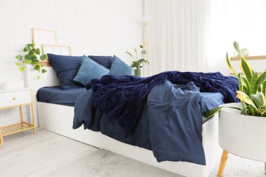 Bed with stylish blue bedding, bedside table and houseplants near white brick wall in bedroom