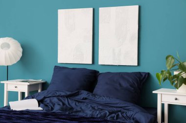 Şık mavi yatak, komodinli komodinli ev bitkisi ve yatak odasındaki duvara asılı resimler.