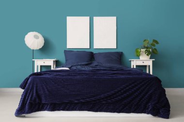 Şık mavi yatak, komodinli komodinli ev bitkisi ve yatak odasındaki duvara asılı resimler.