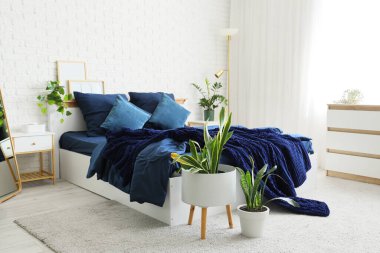 Bed with stylish blue bedding, commode, bedside table and houseplants near white brick wall in bedroom