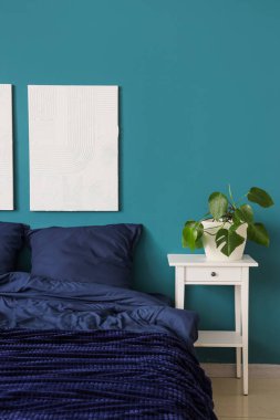 Bed with stylish blue bedding, bedside table, houseplant and hanging paintings on wall in bedroom