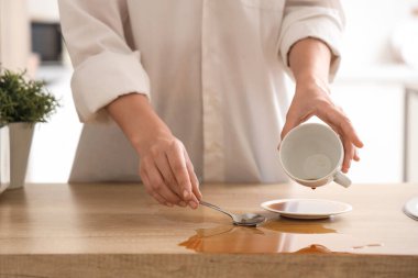 Woman spilling coffee from cup on table in kitchen, closeup