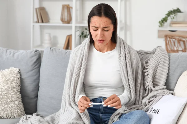 Young woman ill with chickenpox and thermometer sitting on sofa at home