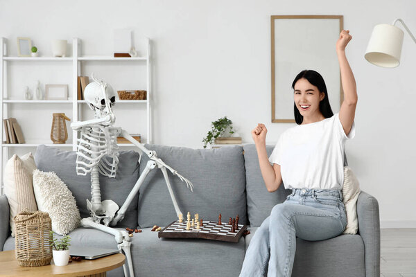 Happy young woman with human skeleton playing chess on sofa at home