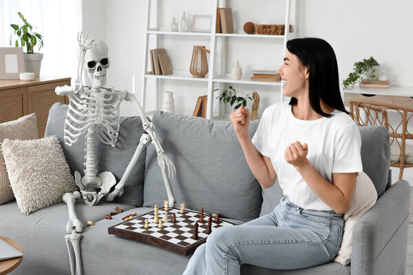 Happy young woman with human skeleton playing chess on sofa at home