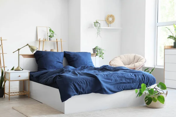 Bed with stylish blue bedding, armchair, shelving unit, bedside table and houseplants near window in bedroom