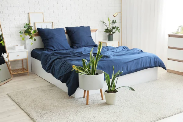Bed with stylish blue bedding, commode, bedside table, lamp and houseplants near white brick wall in bedroom