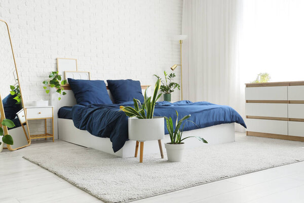 Bed with stylish blue bedding, commode, mirror, bedside table and houseplants near white brick wall in bedroom