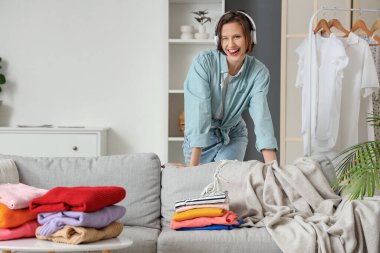 Kulaklıklı genç bir kadın evde farklı giysiler katlıyor.