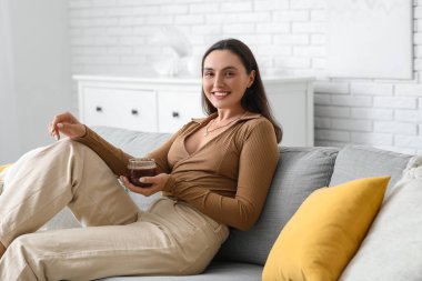 Oturma odasındaki kanepede oturan cam bardak sıcak kakaolu mutlu genç kadın.