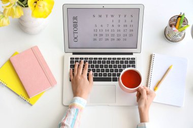 Masada takvim ve çay fincanı olan dizüstü bilgisayarı kullanan kadın eller.