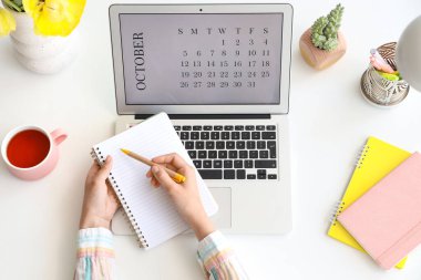 Dizüstü bilgisayarı takvime ve deftere yazı yazan kadın elleri, en üstte görünüm