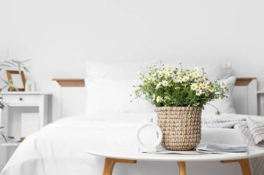 Yatak odasındaki masasında papatya ve dergi olan hasır tencere.