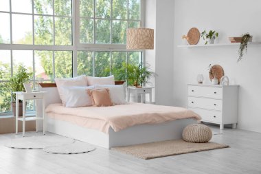 Bed with stylish pink bedding, commode, bedside tables and houseplants near window in bedroom